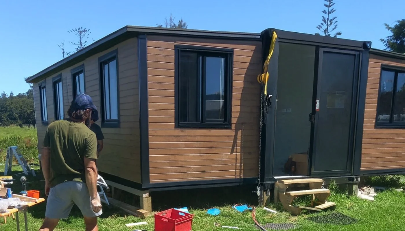A timber-clad expandable home being delivered and set up on a grassy New Zealand site