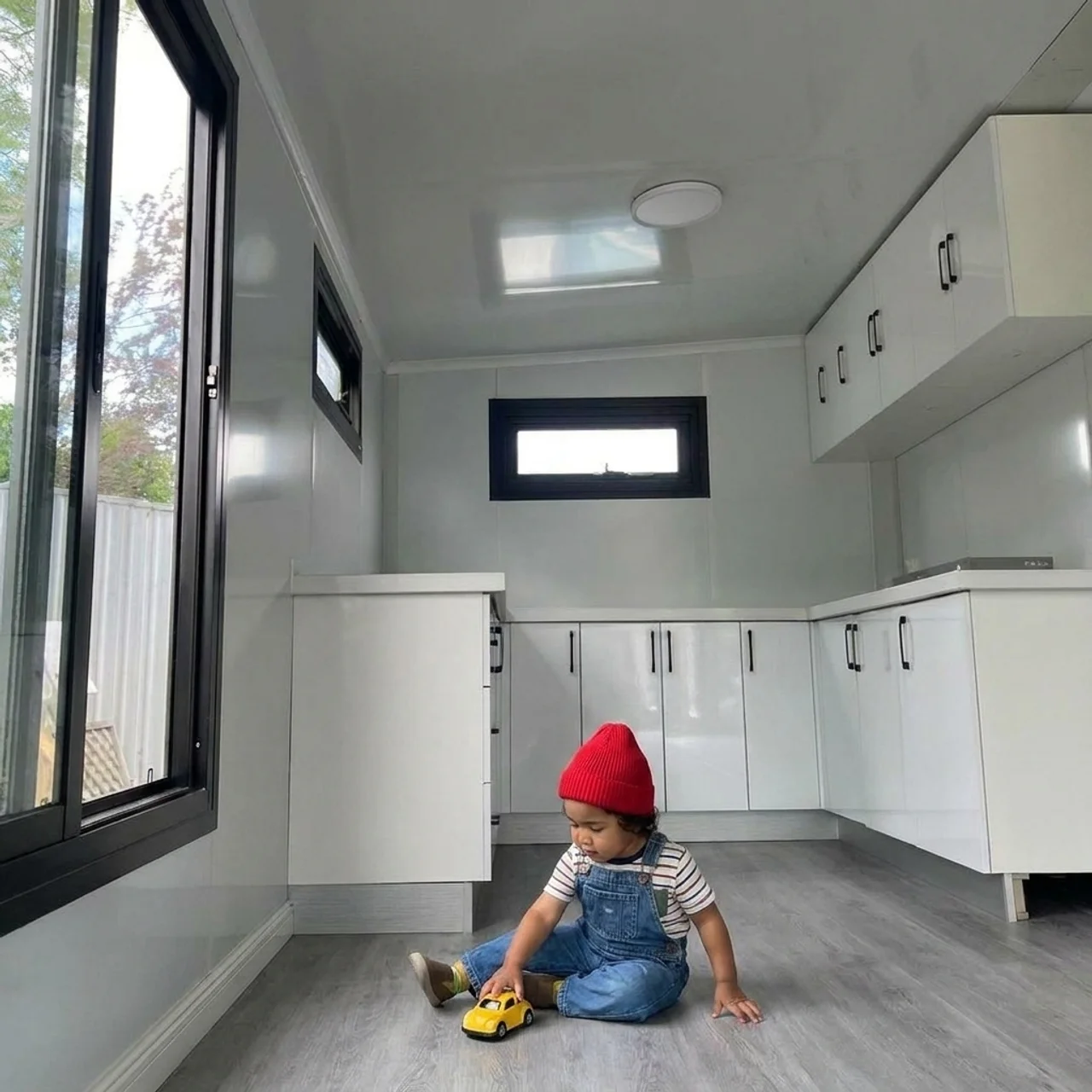 Bright white interior of an expandable tiny home with a toddler in a red beanie playing on the vinyl floor beside the kitchen cabinetry