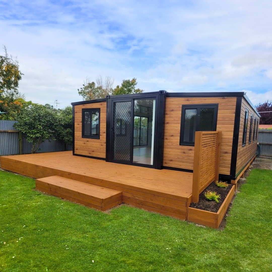 A modern wooden-clad expandable tiny home with a timber deck set on a green lawn in New Zealand