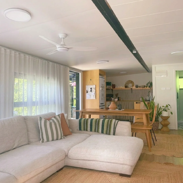 Open-plan living area inside a tiny home with a linen sofa, ceiling fan, timber dining table and compact kitchen