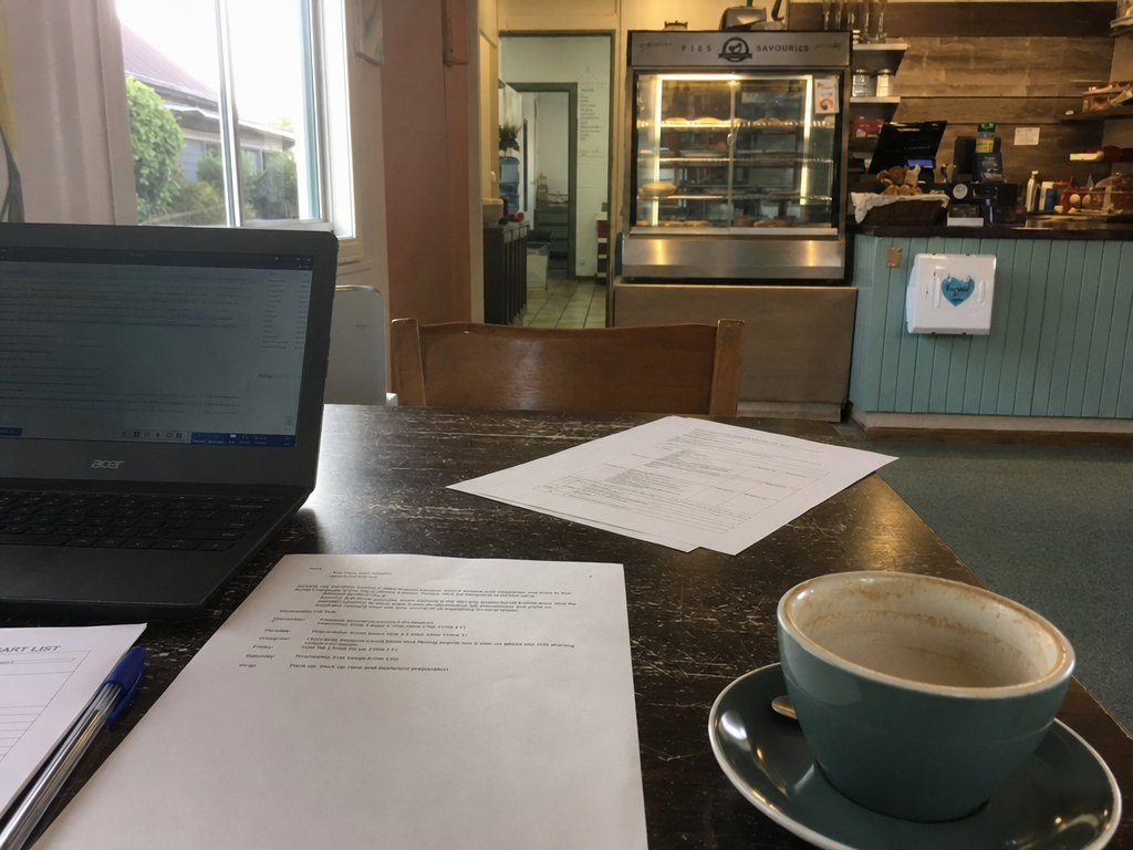 A laptop, papers and an empty coffee cup on a cafe table with a pie warmer and counter in the background